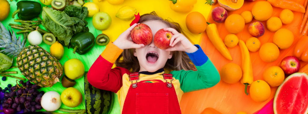 kid surrounded by fruit holding apples in front of eyes
